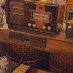Rádio antigo de madeira sobre aparador clássico, ao lado de vasos, livros e objetos decorativos em um ambiente com móveis antigos